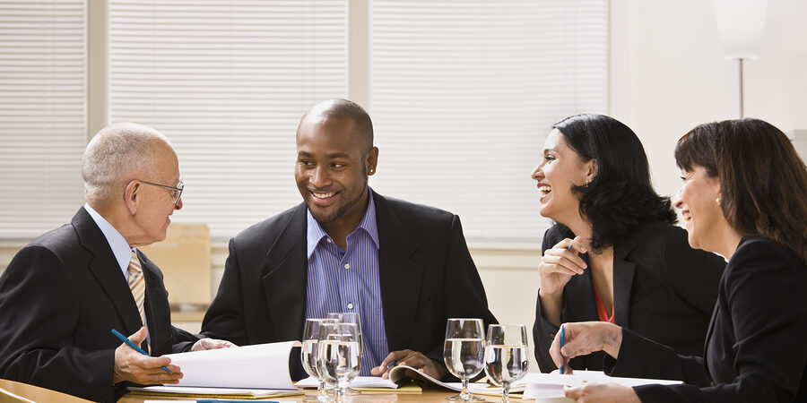 A group of business people are in a meeting in an office.  They are talking and laughing and looking away from the camera.  Horizontally framed shot.