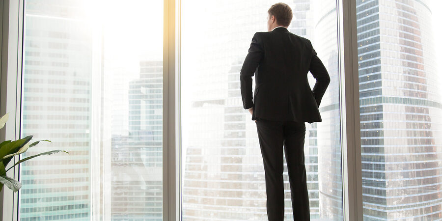 Full length back view of successful businessman in suit standing in office with hands on his waist, CEO looking through window at big city buildings, planning new project, waiting for meeting to start